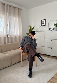 Vyras su akiniais ir berete, sėdintis sukryžiuotomis kojomis ant medinės kėdės minimalistinėje svetainėje su sofa ir baltu spintelėmis.