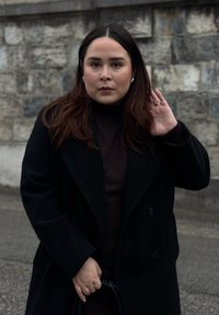 Svart dubbelknäppt ullrock över en mörk polo-tröja, med långt brunt hår. Håller en svart handväska. Stenmur i bakgrunden.