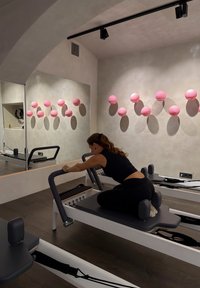 Reformer Pilates-maskine med en sort og hvid ramme, grå måtte og polstrede støtte. Lyserød vægdekoration og et stort spejl i et minimalistisk studio.