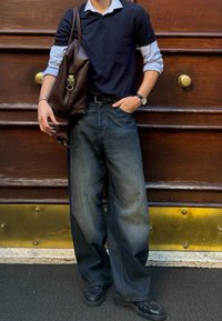 Polo shirt navy sovrapposto a una camicia a maniche lunghe azzurra, jeans scuri larghi, cintura nera, scarpe nere e una borsa a tracolla in pelle marrone.