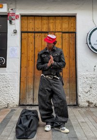 Jachetă din denim negru și pantaloni largi, capac roșu cu logo, adidași albi. Accente din lanț argintiu, curea neagră. Geantă duffle neagră pe sol.