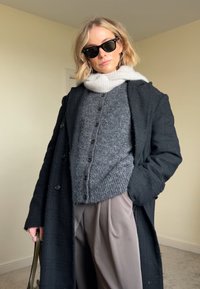 Cappotto nero, cardigan lavorato a maglia grigio con bottoni, sciarpa grigio chiaro, pantaloni taupe e occhiali da sole neri. Texture morbide e look a strati.