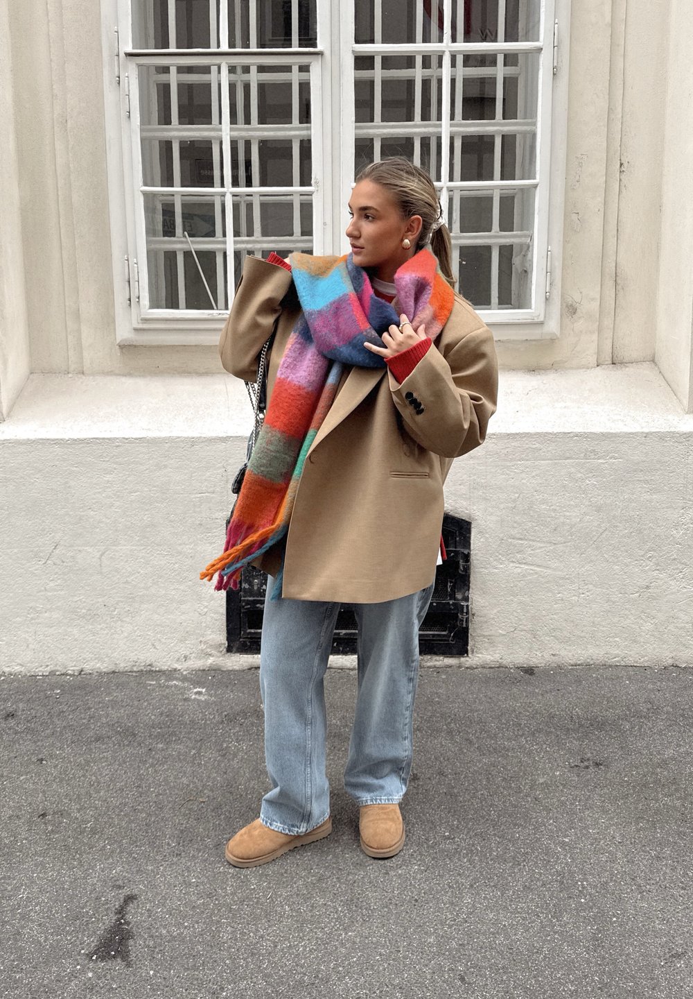 Beige oversized frakk, flerfarget mønstrete ullskjerf, rød genser, lys blå jeans og beige semsket ankelstøvel, mot en hvit vegg.