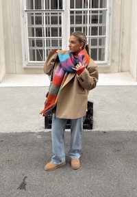 Beige oversized frakk, flerfarget mønstrete ullskjerf, rød genser, lys blå jeans og beige semsket ankelstøvel, mot en hvit vegg.