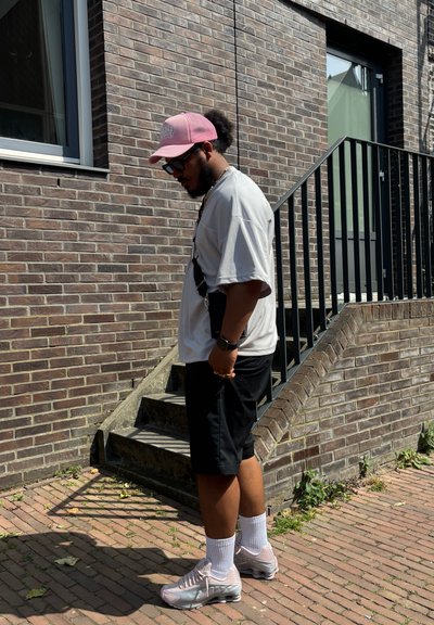 El atuendo informal consiste en una camiseta gris suelta, pantalones cortos negros, una gorra rosa y zapatillas ligeras rosas. De pie al lado de una pared de ladrillos y escaleras.