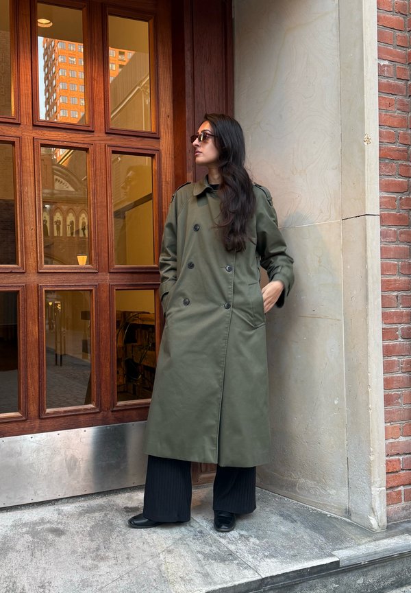Una mujer con gafas de sol y un abrigo largo de color verde oliva está de pie junto a una ventana de madera y mira hacia un lado. Lleva pantalones oscuros y zapatos.