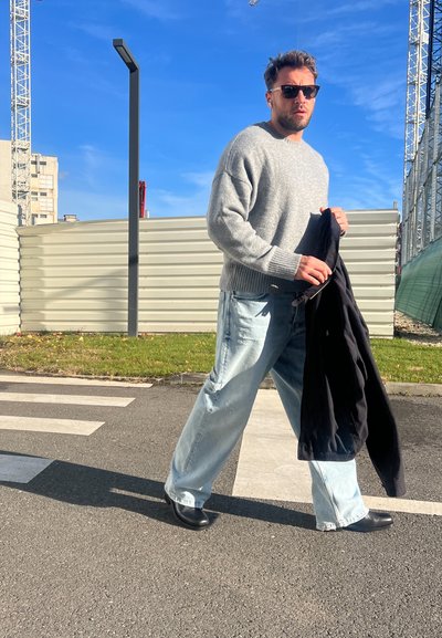 Suéter gris de punto, jeans sueltos azul claro, zapatos negros. Sostenido una chaqueta negra, caminando por una calle con paso de peatones, cielo azul claro.