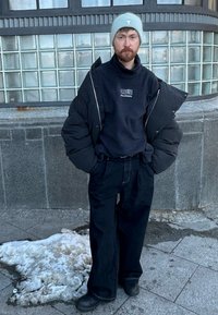 Homme debout sur un trottoir urbain enneigé, portant une doudoune noire sur un sweat-shirt noir, un pantalon large noir, des bottes et un bonnet gris clair.