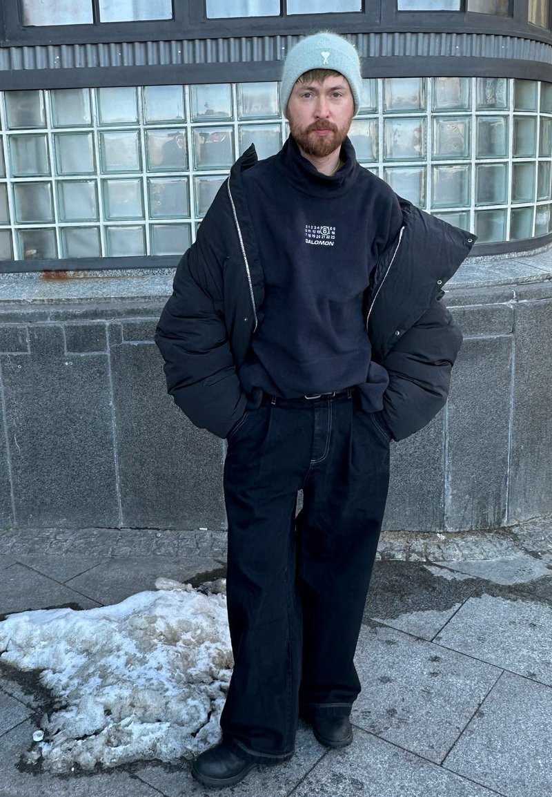 Homme debout sur un trottoir urbain enneigé, portant une doudoune noire sur un sweat-shirt noir, un pantalon large noir, des bottes et un bonnet gris clair.