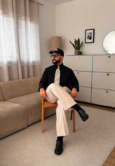 Hombre con gafas, camisa negra, corbata a rayas y gorra sentado con las piernas cruzadas en una silla de madera en una sala de estar moderna con sofá beige y armario.