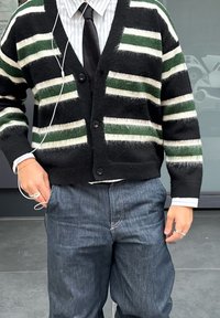 Cardigan noir avec des rayures horizontales vertes et blanches, doté d'un col en V, de boutons et de poignets côtelés, porté par-dessus une chemise à col et un pantalon.
