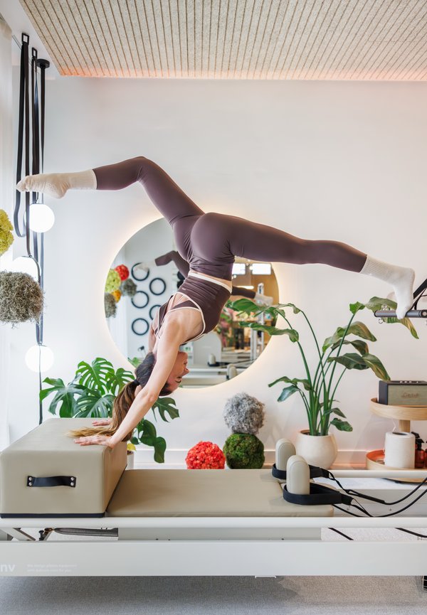 Kobieta w sportowym stroju wykonująca stanie na rękach z rozkrokiem na maszynie do Pilates w jasnym, pełnym roślin studio.