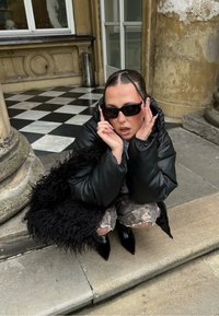 Musta puffertakki, teksturoitu musta tekoturkislaukku, kuviolliset shortsit ja terävät mustat saappaat. Aurinkolasit päässä; kädet kehystävät kasvoja.