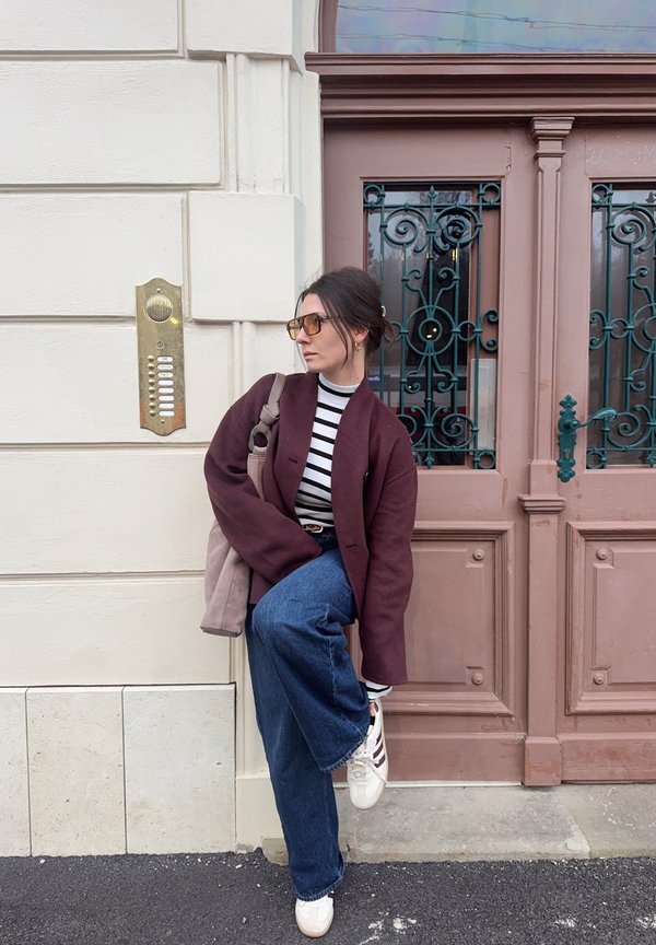 Γυναίκα που ρυθμίζει το λευκό αθλητικό παπούτσι, φοράει μπορντό παλτό, ριγέ μπλούζα, φαρδιά μπλε τζιν, γυαλιά ηλίου, στέκεται δίπλα σε μπεζ τοίχο και περίτεχνη ξύλινη πόρτα.