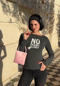 Femme aux cheveux courts noirs avec un bandeau, portant un t-shirt rayé avec l'inscription « NO DRAMA ALLOWED », tenant un sac rose, levant l'index en plein air sous la lumière du soleil.