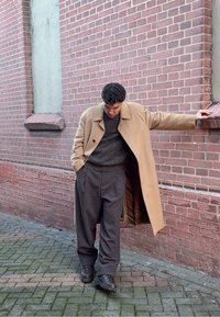 Manteau beige, pull gris texturé et pantalon ample foncé. Le mannequin s'appuie contre un mur de briques. Chaussures noires à semelles épaisses.