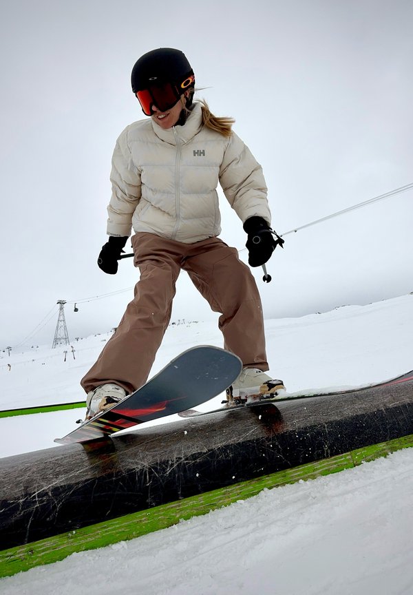 Esquiando en un riel negro, una persona viste una chaqueta de plumas beige, pantalones marrones y un casco negro con detalles naranjas, contra un fondo nevado.