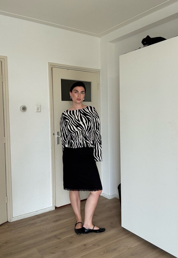 Kvinde stående indendørs iført en sort og hvid zebra-print top, sort nederdel og sorte balletflade sko på et trægulv.