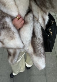 Personne portant un manteau en fourrure blanc et gris, pantalon de couleur crème, chaussures noires, bague en or, tenant un sac à main en cuir noir avec des détails en chaîne dorée.