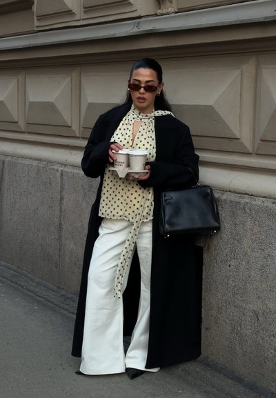 Mujer con abrigo negro y blusa de lunares sosteniendo dos tazas de café, de pie frente a una pared decorativa de piedra en una calle de la ciudad.