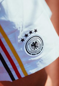 Bela kratka hlače z okroglo črno oznako, na kateri je dvojna glava orla in besedilo "Deutscher Fussball-Bund", dopolnjena s črnimi, rdečimi in rumenimi črtami.