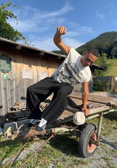 Pantalones negros a rayas, camiseta sin mangas blanca con gráfico, zapatillas negras. Sentado en un carro de madera, sosteniendo una gorra de béisbol color crema. Paisaje verde.