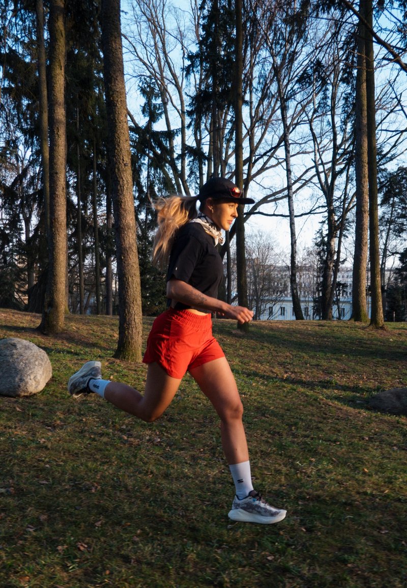 Femme portant une chemise noire, un short rouge et une casquette, courant à travers un parc avec des arbres sans feuilles et des rochers sur l'herbe en plein jour.