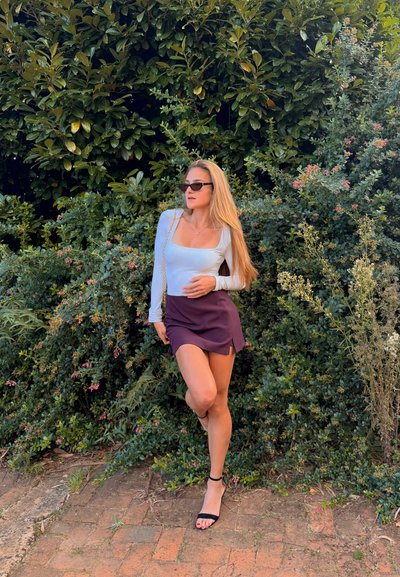 Mujer con top de manga larga blanco, falda morada y sandalias negras posando frente a una densa vegetación verde en un camino de ladrillos.