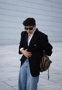Μαύρο σακάκι με καρφίτσα πέτου, λευκό πουκάμισο με φουσκωμένα μανίκια και φαρδιά τζιν παντελόνια σε μπλε απόχρωση. Κρατώντας μια καφέ τσάντα με κορδόνι, φοράει σκούρα γυαλιά ηλίου.