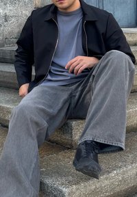 Čierna zipsová bunda cez sivé tričko, skombinovaná s vyblednutými širokými džínsami v sivej farbe a čiernymi topánkami, sediac na kamenných schodoch.