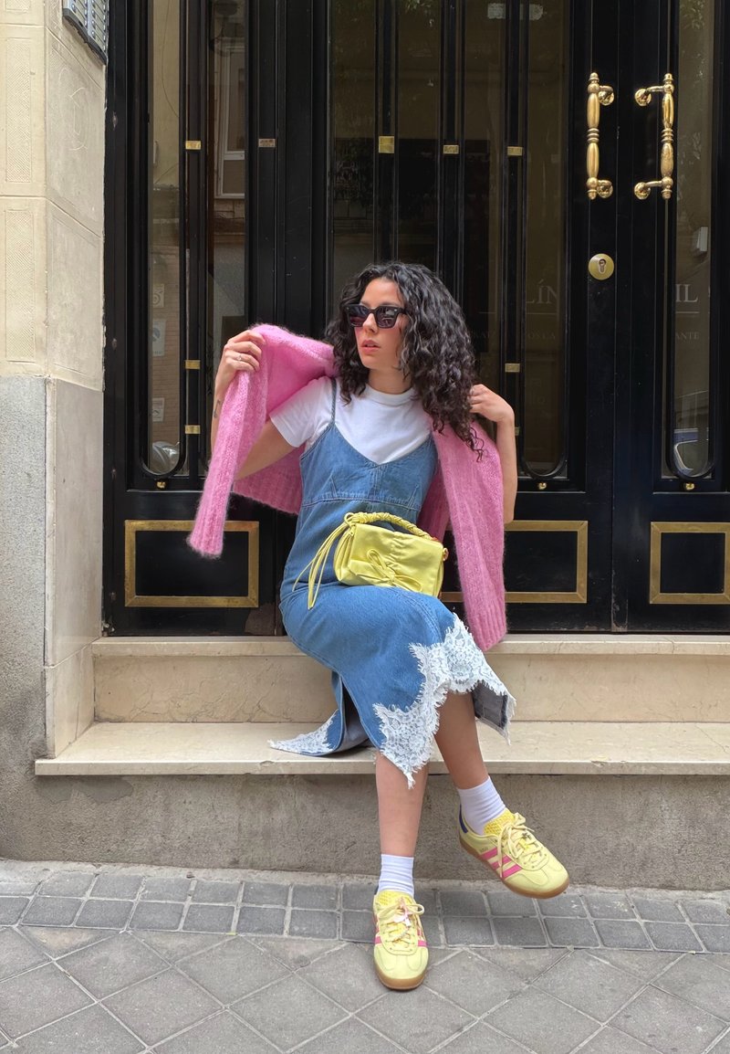 Femme aux cheveux bouclés portant des lunettes de soleil, un cardigan rose sur une chemise blanche et une robe bleue ornée de dentelle, assise sur une marche avec un sac jaune et des baskets.