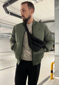 Gestepptes olivgrünes Bomberjackett, grauer Pullover und schwarze Umhängetasche. Glatte Texturen mit einer Reißverschlusstasche an der Tasche. Innenraum.