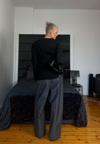 Femme blonde vêtue d'un haut noir et d'un pantalon large gris, debout de dos dans une chambre avec une couette noire et un sol en bois.