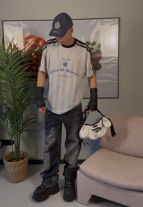 Gestreiftes T-Shirt in Schwarz und Weiß mit blauer Schrift, schwarze oversized Jeans und klobige schwarze Schuhe, in der Hand eine weiße Handtasche mit schwarzem Riemen.