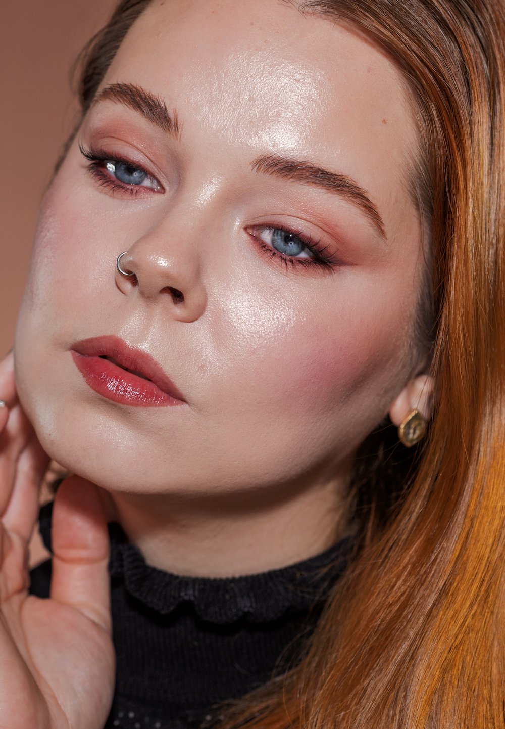 Nærbilde av et ansikt med lys hud, blå øyne og langt rødbrunt hår. Kjennemerker inkluderer en sølvnese-ring og gull øredobber.
