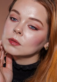 Nærbilde av et ansikt med lys hud, blå øyne og langt rødbrunt hår. Kjennemerker inkluderer en sølvnese-ring og gull øredobber.