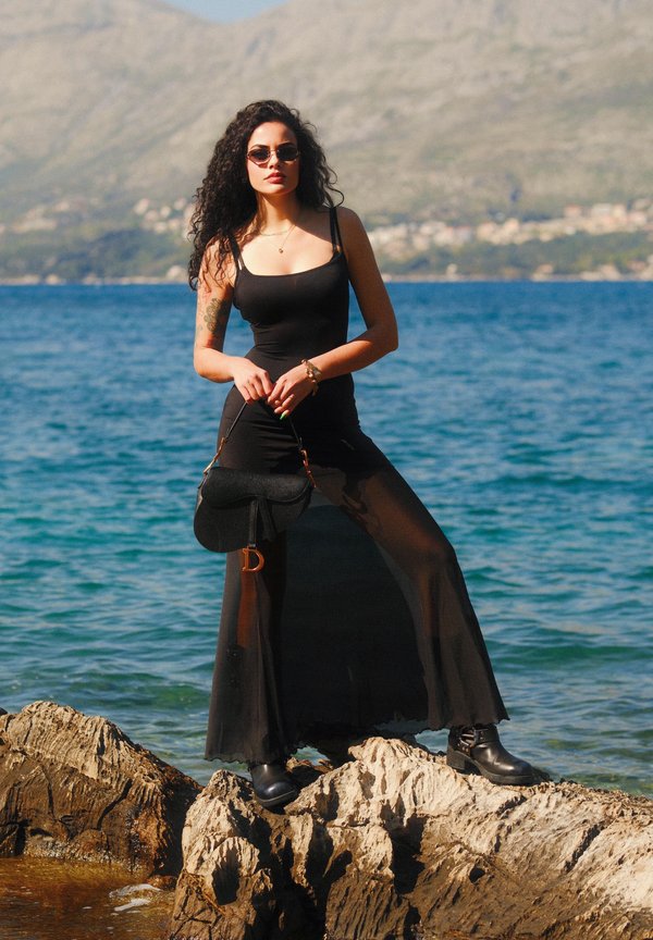 Abito aderente nero con porzioni di gonna trasparente, abbinato a stivaletti neri alla caviglia. Gli accessori includono una piccola borsa nera e occhiali da sole. Sfondo con una scogliera rocciosa.