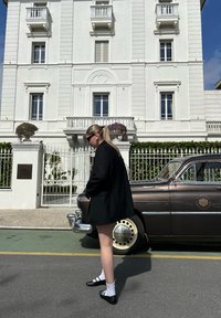 Blazer nero, camicia bianca e scarpe nere con strisce bianche. Auto d'epoca marrone con dettagli cromati parcheggiata su una strada di fronte a un edificio bianco.
