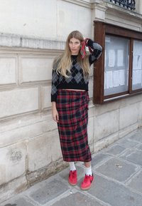 Black argyle jumper, red tartan wrap skirt, and bright red trainers. Long, wavy hair adorned with a red scarf accessory. Urban backdrop.