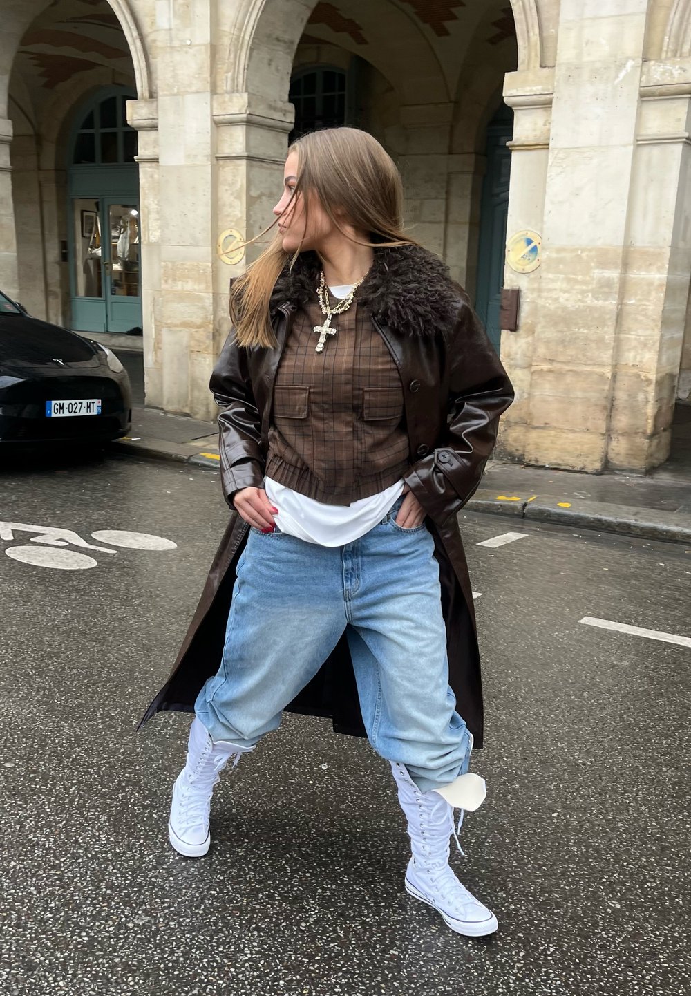 Jeune femme debout dans une rue mouillée, portant un manteau marron foncé, une chemise à carreaux, un jean bleu clair, des baskets montantes blanches et des colliers superposés avec une croix.