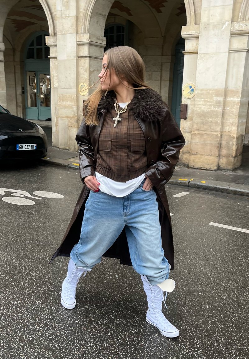 Jeune femme debout dans une rue mouillée, portant un manteau marron foncé, une chemise à carreaux, un jean bleu clair, des baskets montantes blanches et des colliers superposés avec une croix.