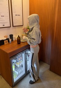 Personne en sweat à capuche gris et pantalon tenant un petit verre au comptoir en bois avec un mini-réfrigérateur et des menus de boissons accrochés au mur.