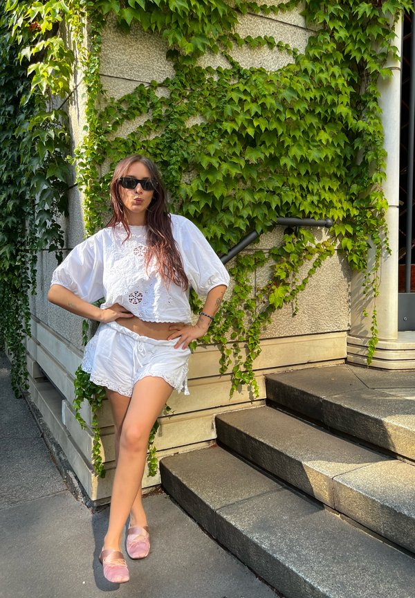Blusa bianca a maniche corte con dettagli in pizzo abbinata a pantaloni corti bianchi. La persona indossa delle scarpe flat rosa e occhiali da sole, posata accanto a edera verde.