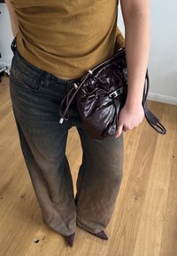 Brązowa skórzana torebka o marszczonym wzorze i srebrnym wykończeniu, zestawiona z ciemnymi jeansami z wysokim stanem i spiczastymi burgundowymi butami na drewnianej podłodze.