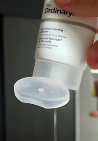 Die Tube des The Ordinary Glucoside Foaming Cleanser wird kopfüber gehalten und gibt eine klare Flüssigkeit aus dem offenen Deckel ab.
