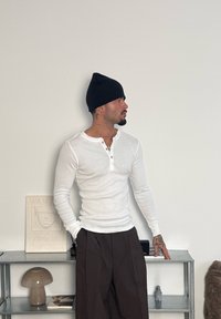 Chemise Henley blanche à manches longues avec boutons, associée à un pantalon large brun foncé. Bonnet noir et tatouages visibles sur le bras.
