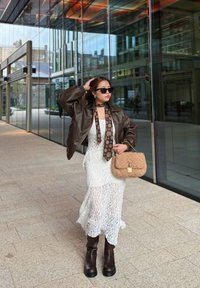 Kaffe kleur leren cropped jack over een witte kanten jurk met een hoge split. Zwarte zonnebril, een patroontie en een gestructureerde beige handtas. Bruine kniehoge laarzen.