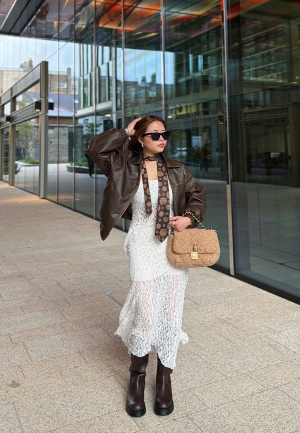Chaqueta de cuero marrón corta sobre un vestido de encaje blanco con una gran abertura. Gafas de sol negras, una corbata con estampado y un bolso de mano texturizado en beige. Botas marrones hasta la rodilla.