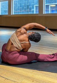 Une personne portant un brassard de sport beige et des leggings bordeaux effectue un étirement latéral sur un tapis dans un espace intérieur bien éclairé.