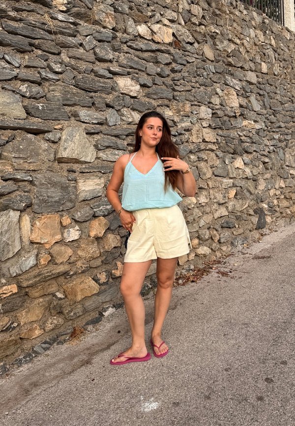 Jeune femme portant un haut bleu clair, un short crème et des tongs roses, debout sur un chemin pavé à côté d'un mur en pierre rugueuse.